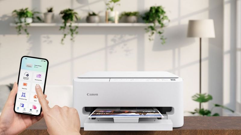 A Canon PIXMA printer, white in colour, prints a sheet of A4 paper in a home setting, and a hand holding a smartphone.