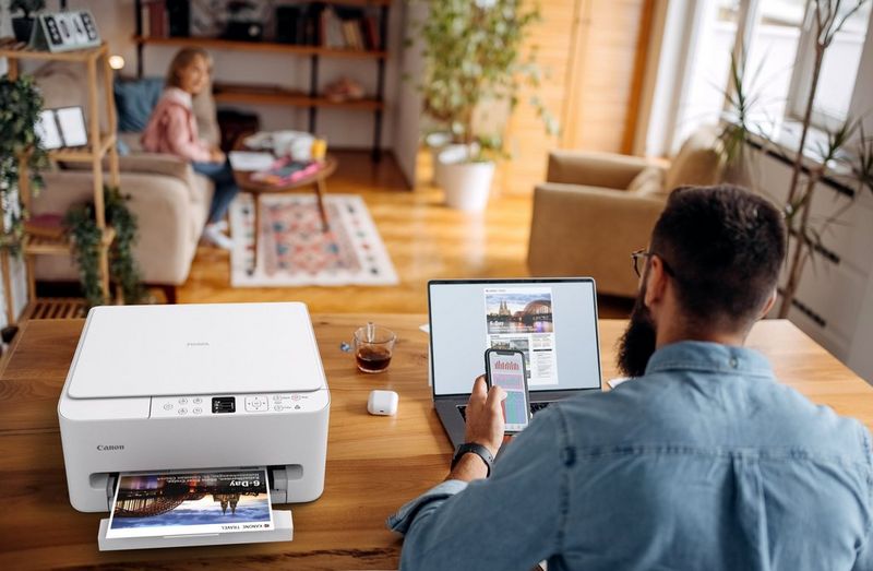 גבר מחזיק סמארטפון ויושב ליד שולחן בבית מודרני, כשמדפסת Canon אלחוטית מדפיסה תמונה שמופיעה גם על מסך הלפטופ שלצידה.