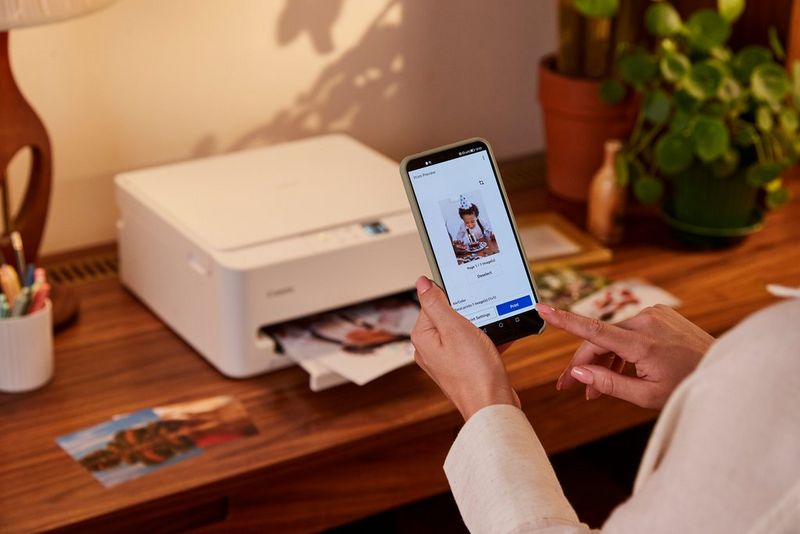 Žena drží mobilný telefón, na ktorom vidieť fotografiu v aplikácii Canon PRINT. Tlačiareň Canon PIXMA sa nachádza na drevenom povrchu v pozadí.