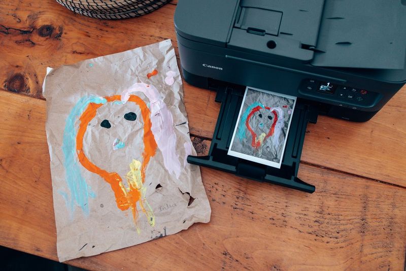 A black Canon printer prints a child's painting on Canon Magnetic Photo Paper. The original piece of art is on the table next to the printer.