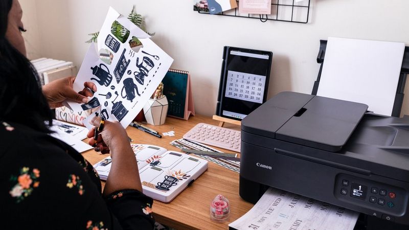 Een vrouw snijdt Creative Park-sjablonen uit terwijl ze achter een houten bureau zit. Op het bureau staat een Canon PIXMA-printer en liggen hobbymaterialen.