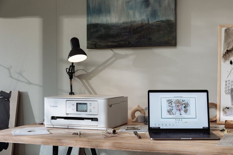 A laptop and a Canon PIXMA printer sit on a wooden table in a home, with a black lamp overlooking the printer.