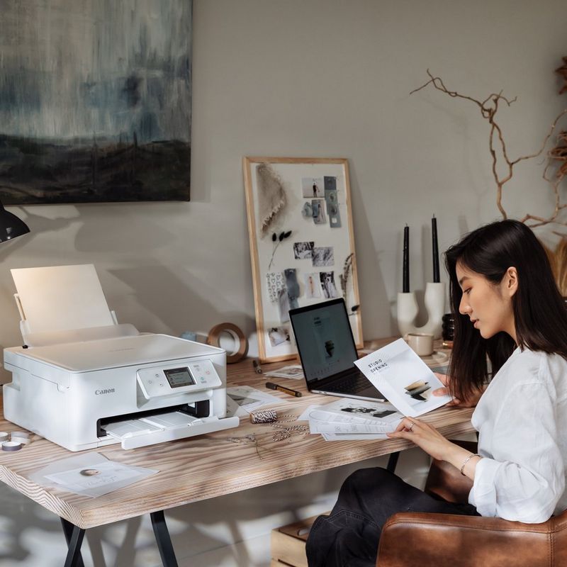 Ženska gleda kup papirjev, tiskalnik Canon PIXMA in druge pisarniške potrebščine pa so na mizi.