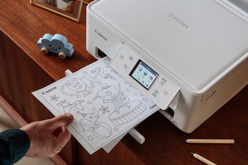 Op een houten dressoir staat een Canon PIXMA-printer. Een hand pakt een print met kleurontwerpen uit de lade.