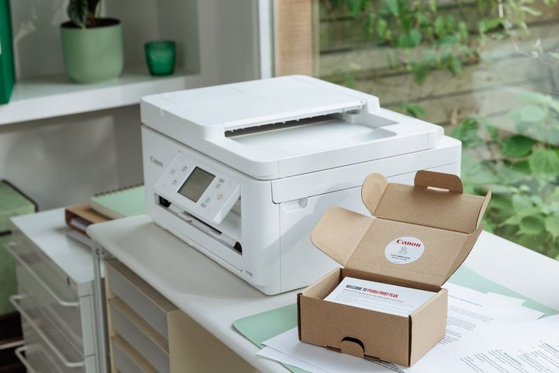 Una confezione Canon PIXMA Print Plan accanto a una stampante Canon PIXMA in un ufficio domestico, con vista sul verde visibile attraverso la finestra sullo sfondo.