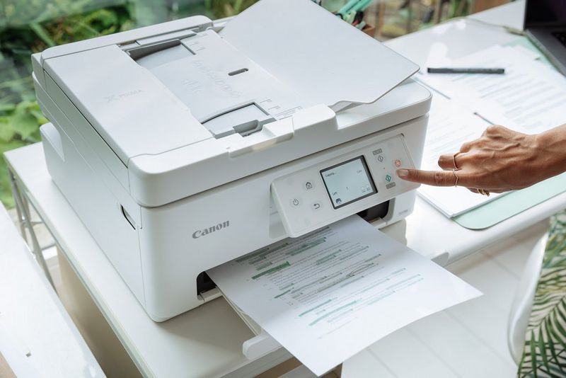 A user's hand presses a button on the control panel of a Canon PIXMA printer sitting on am office desk.