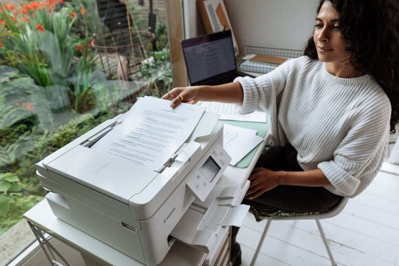 A woman places a pile of documents in the document feed on top of a Canon PIXMA printer on the desk beside her.