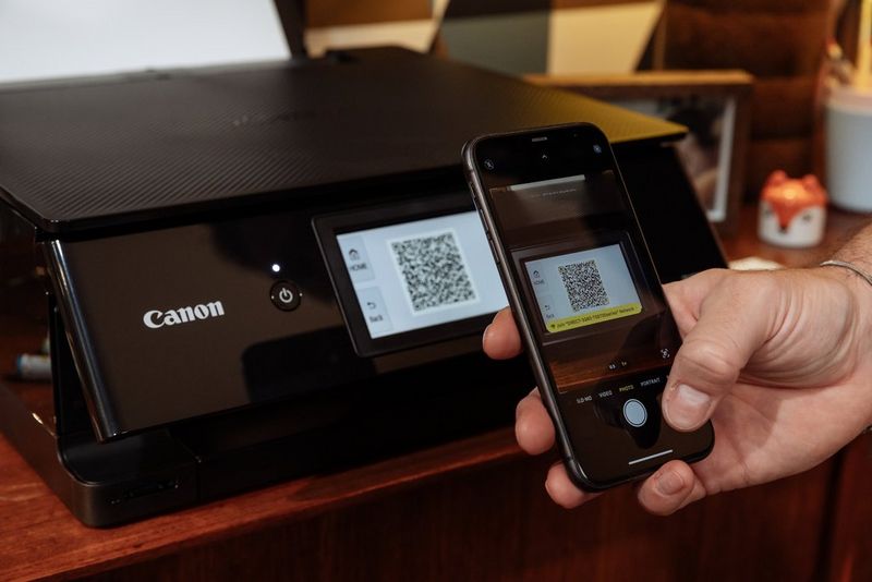 Op een plank staat een Canon PIXMA-printer. Er zit blanco papier in de achterste lade en het kleurentouchscreen licht op.