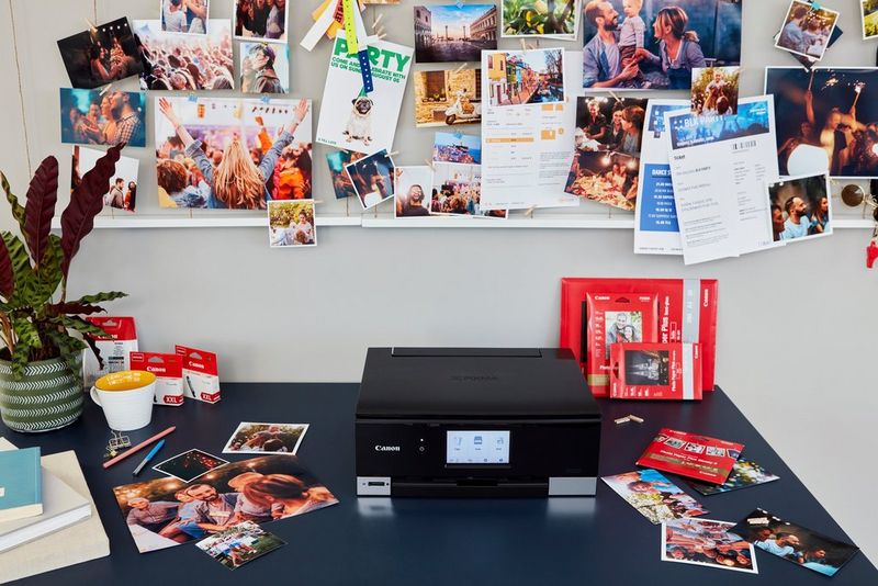 Принтер Canon PIXMA, фотографии разных размеров, пачки бумаги и чернила на столе. На стене сзади — пробковая доска с закрепленными на ней распечатками разного типа и размера.