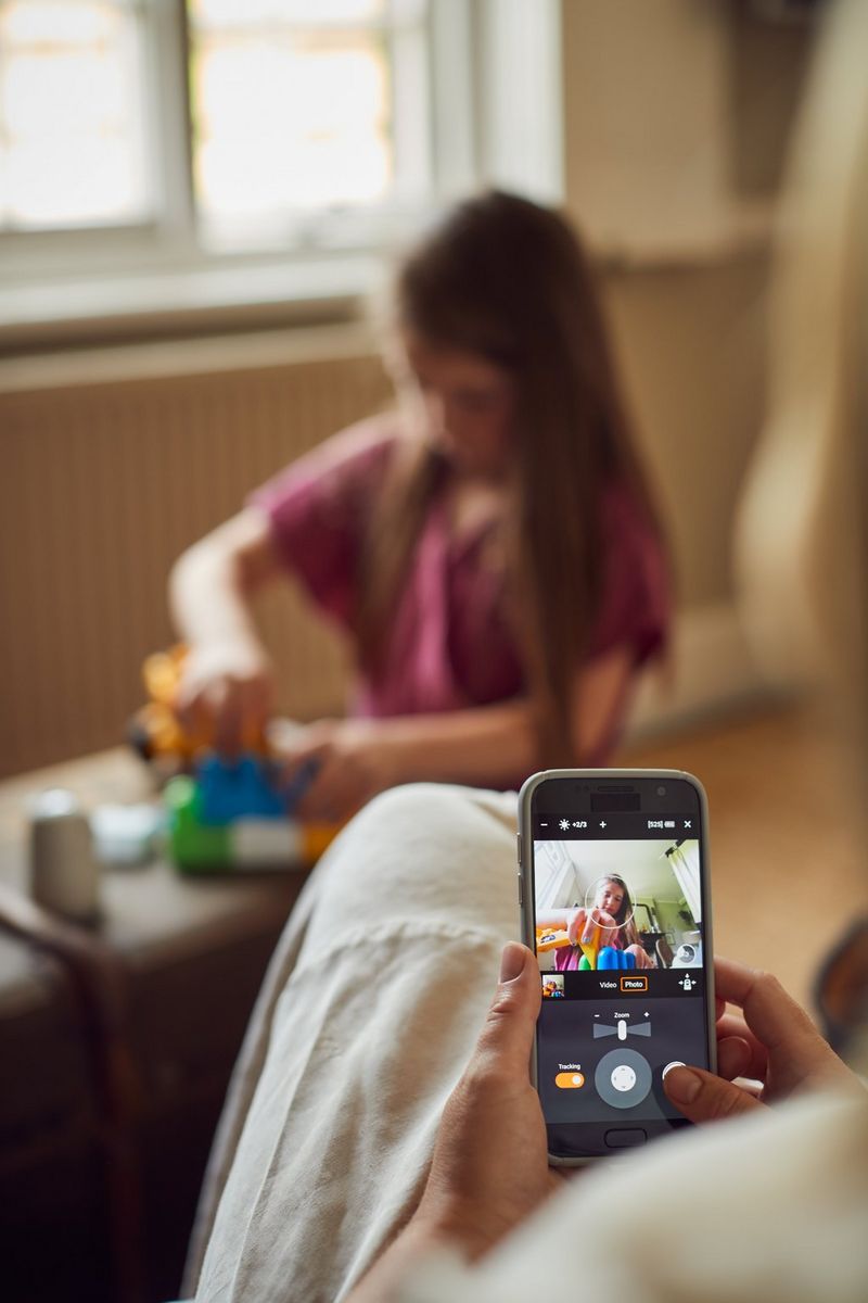 A girl kneels at a low table playing with toys, the Canon PowerShot PX in front of her. Her mother is viewing the feed from the camera on her phone, and the photo is visible on the screen as she adjusts the shot.