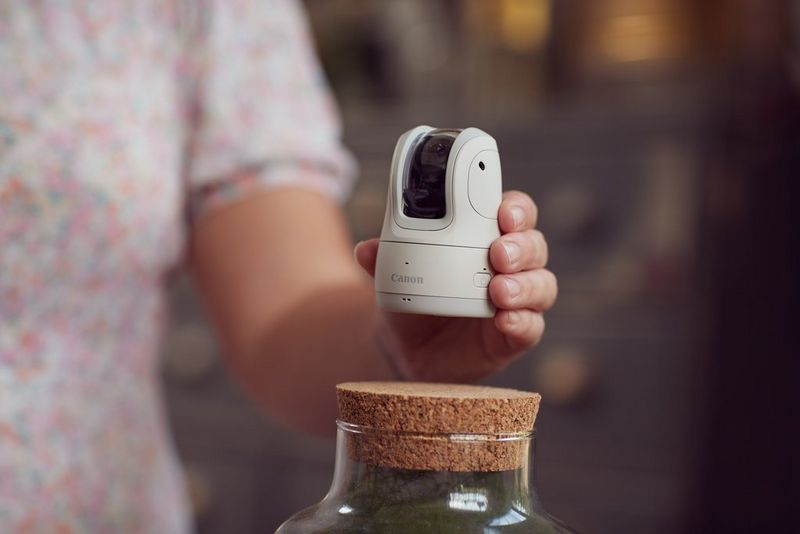 Белая камера Canon PowerShot PX в руках женщины, которая ставит ее на пробковую крышку, находясь на открытом воздухе.