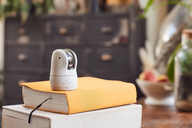 Белая камера Canon PowerShot PX на небольшой стопке книг на деревянном столе.
