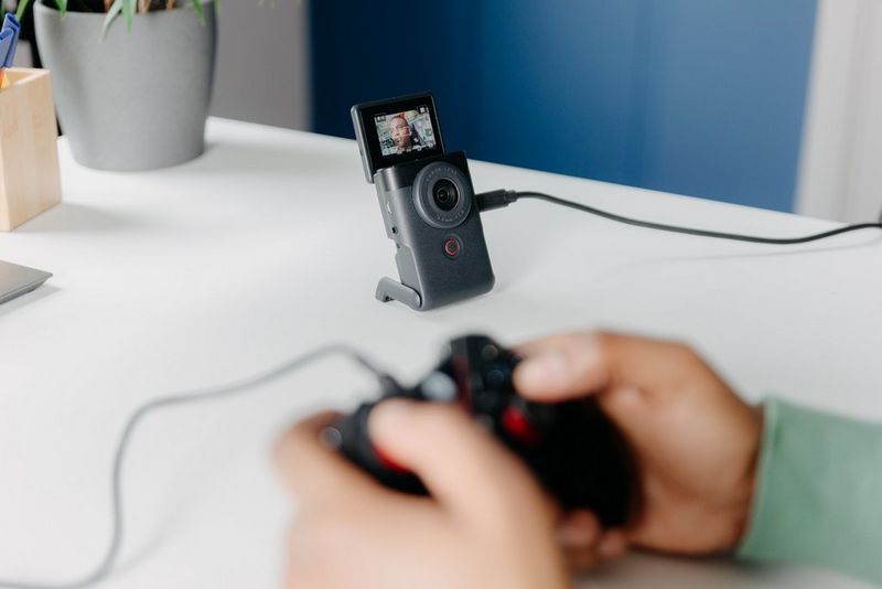 A Canon PowerShot V10, standing on its built-in stand, records a gamer, whose hands using a game controller are visible in the foreground.