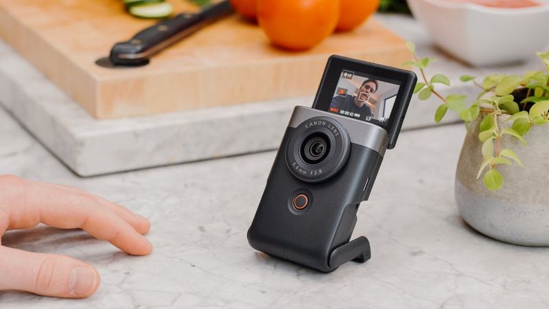 A Canon PowerShot V10 stands on a kitchen worktop with a chopping board in the background and a vlogger's hand resting on the surface in front of it.