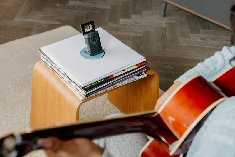 Kamera Canon PowerShot V10 na hromádce alb na postranním stolku natáčí člověka hrajícího na kytaru.