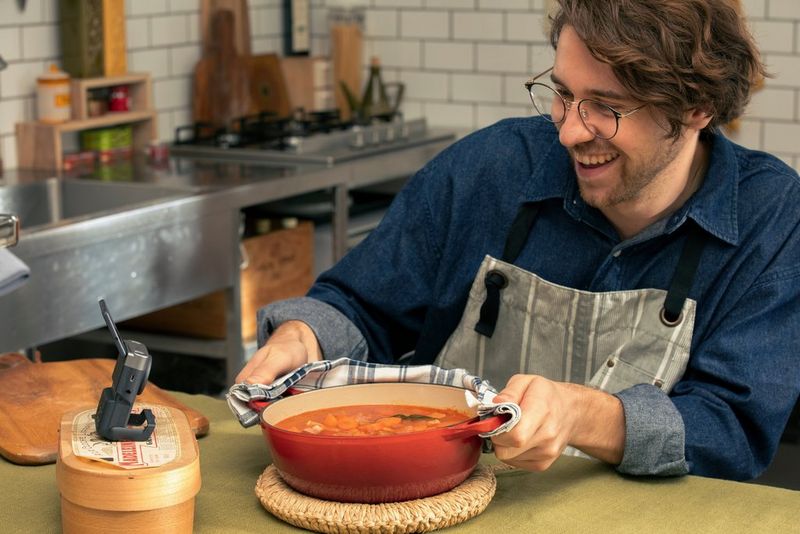 A man in an apron, smiling, holds a red pot of soup with a PowerShot V10 on a stand recording him in a kitchen.