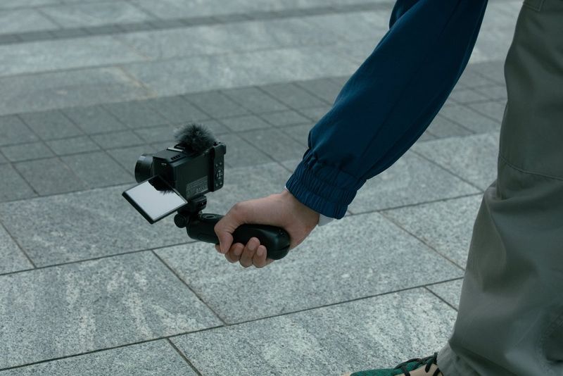 Using a tripod grip, a user holds a Canon PowerShot V1 camera at a low angle with the vari-angle screen angled upwards, suitable for street photography. 