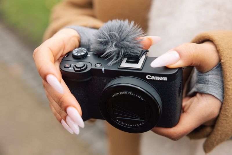 Canon PowerShot V1 v rokah uporabnika na prostem, s kosmato vetrno zaščito, pritrjeno na vrh fotoaparata, da zmanjša šum vetra.