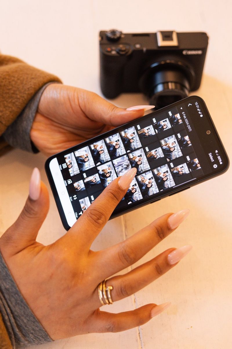 Uživatel drží v ruce chytrý telefon s aplikací Canon Camera Connect, na němž je vidět obrazovku plnou miniatur snímků. Videokamera Canon PowerShot V1 je umístěna níže na stole.