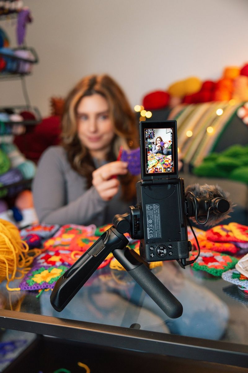 Žena drží v ruce pletení a přitom se natáčí videokamerou Canon PowerShot V1 orientovanou svisle pomocí gripu s funkcí stativu HG-100TBR na skleněném stole.