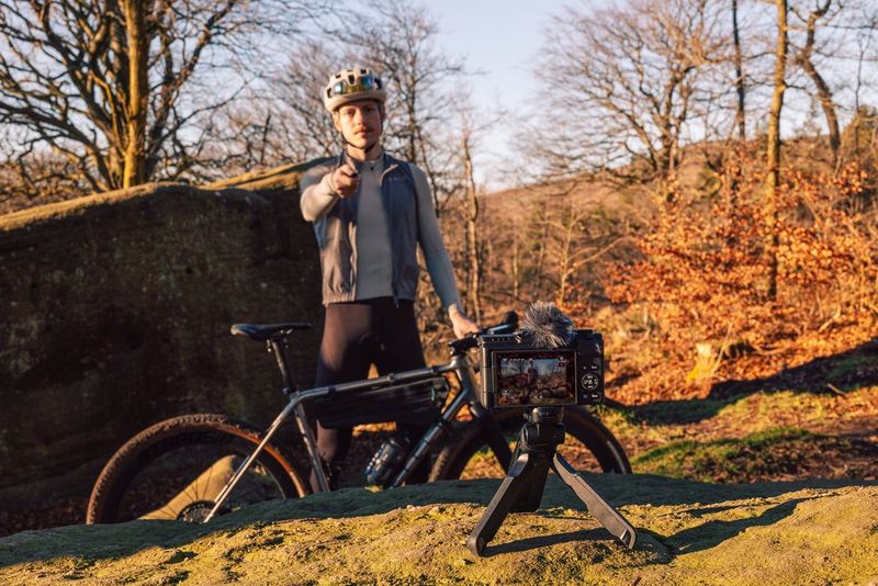 A man wearing a cycling helmet stands behind a bike, gesturing towards a Canon PowerShot V1 on a tripod facing him.