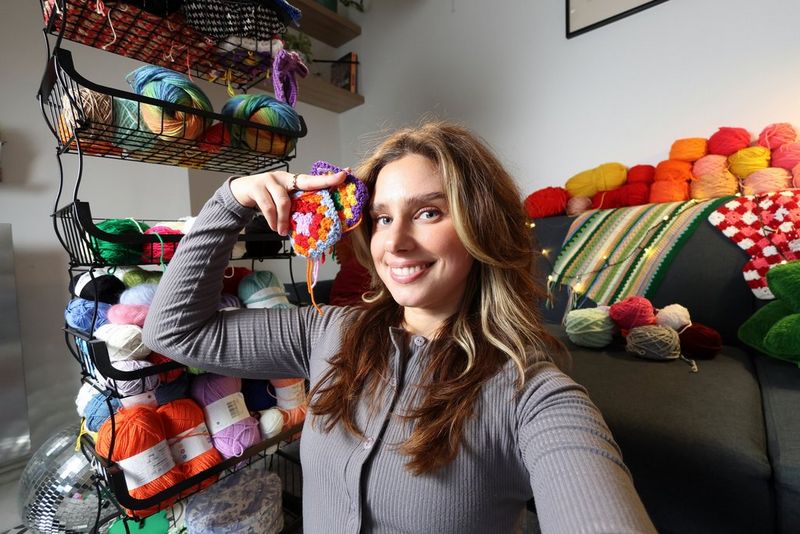 A woman holds up a crochet sample while filming herself with one hand. A wire stand in the background holds balls of wool. 