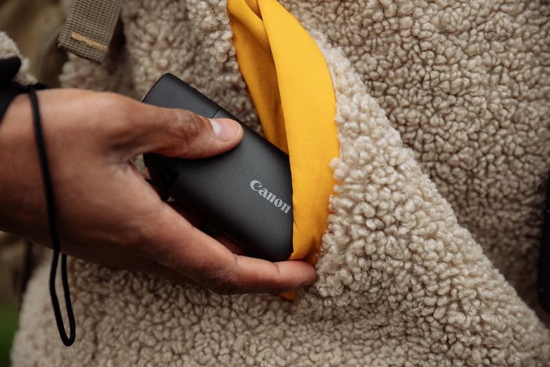 A hand placing a Canon PowerShot ZOOM into the pocket of a fluffy cream and yellow jacket.