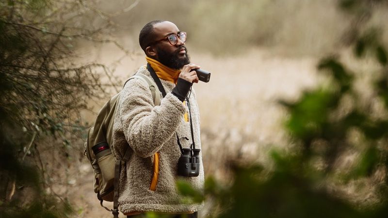 A man stands among foliage in a bird reserve, holding a Canon PowerShot Zoom in one hand and wearing a pair of Canon binoculars around his neck.