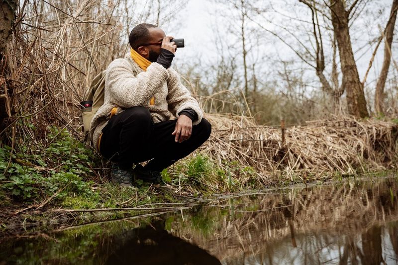 A man crouches at the edge of a stream, holding a Canon PowerShot ZOOM up to his eye.