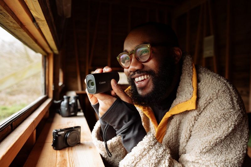A smiling man brings a Canon PowerShot ZOOM up to his face while looking through the window of a bird hide.