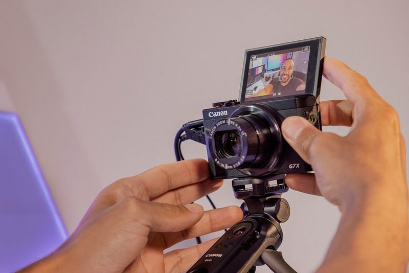 Изображены руки, регулирующие положение ЖК-экрана камеры Canon PowerShot G7 X Mark III на штативе. Человек поворачивает его на себя, чтобы видеть свое изображение во время съемки.