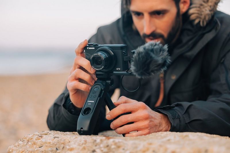 A man leans on a rock while holding a Canon PowerShot G7 X Mark III with a mic attached.