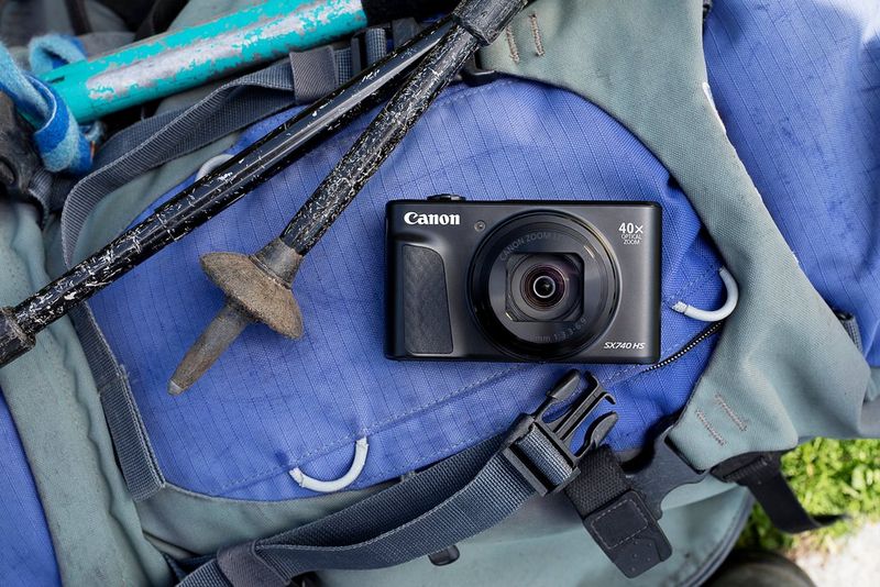 תרמיל גב בצבע סגול ואפור מונח על הרצפה, ליד מוטות הליכה, ומעל שניהם מצלמה מדגם Canon PowerShot SX740 HS LITE EDITION.