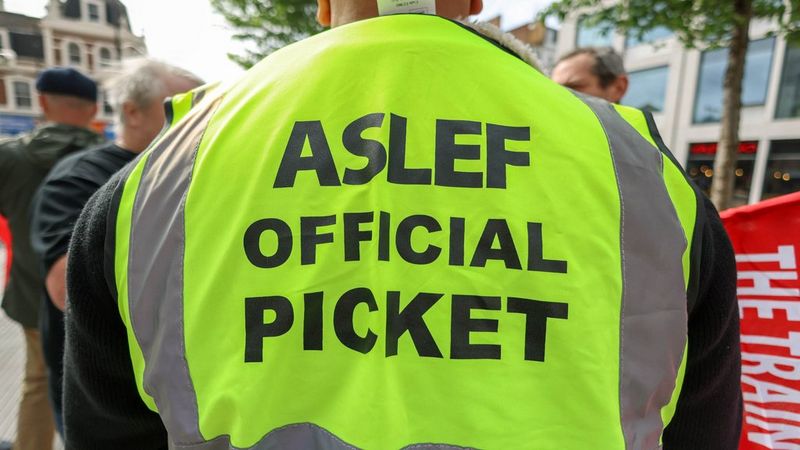 The back of a man wearing a hi-vis fluorescent yellow vest with the words ‘ASLEF OFFICAL PICKET’ printed on it. © Martin Wheatley