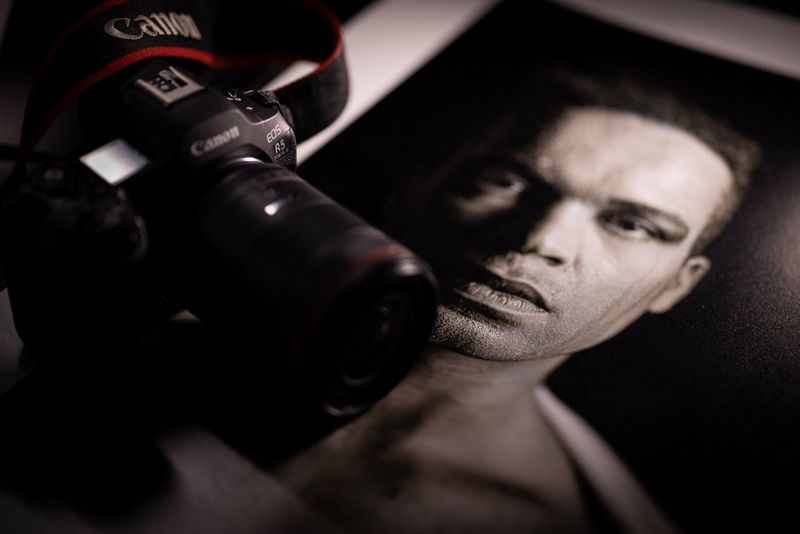 На черно-белом портрете крупным планом — лицо мужчины, по которому струится пот; на снимке лежит камера Canon EOS R5 Mark II.