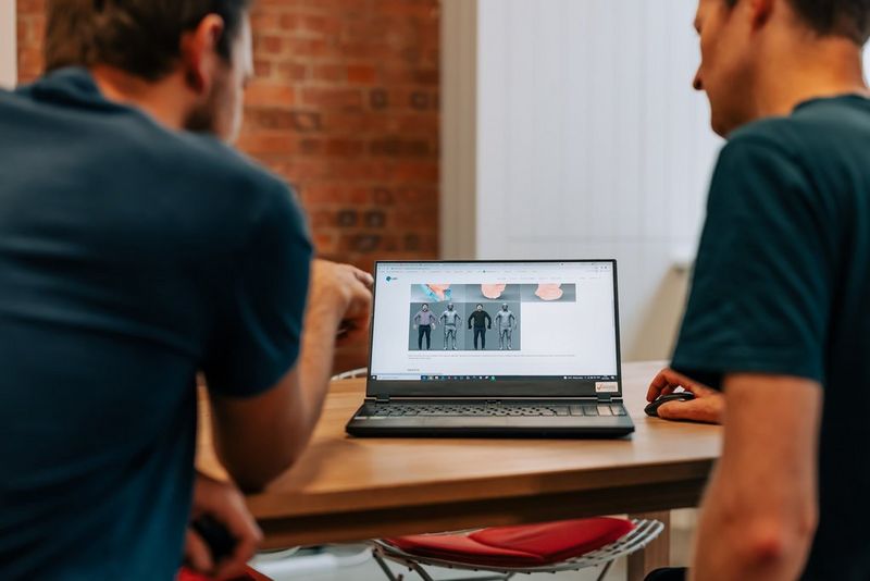 Due uomini seduti a un tavolo guardano lo schermo di un laptop che mostra dei modelli 3D di figure umane.