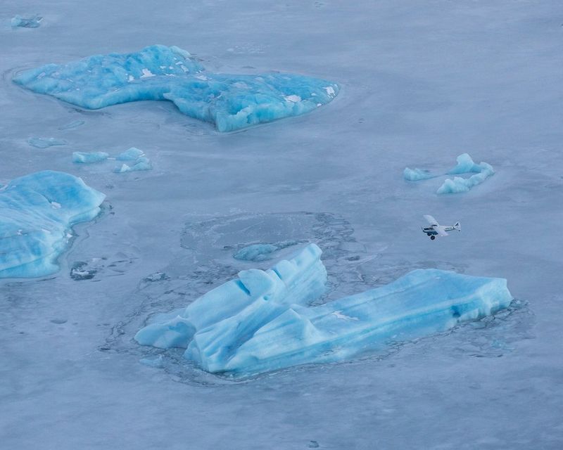 An aerial photograph of light blue glaciers surrounded by ice, with a bush plane flying low above them. 