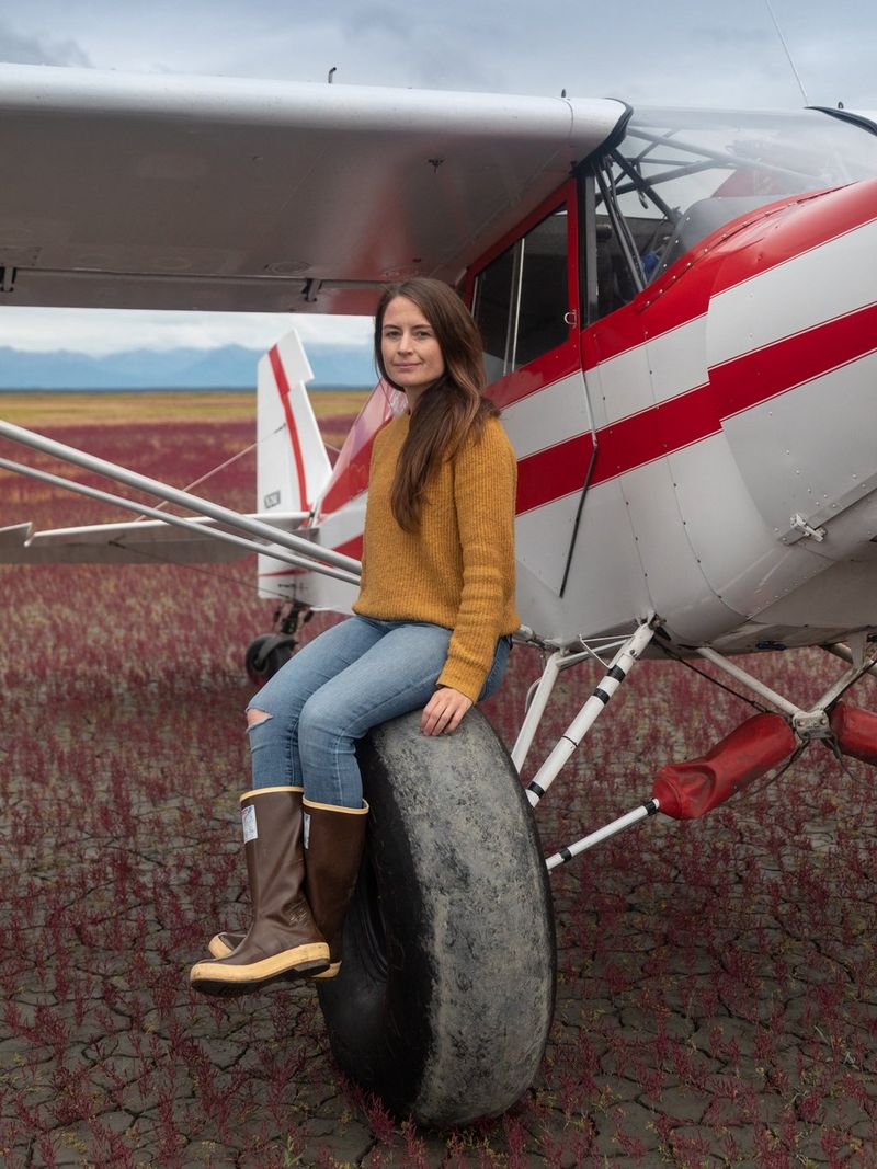Photographer Acacia Johnson, wearing jeans and a thick yellow jumper, sits on the large wheel of an Alaskan bush plane.