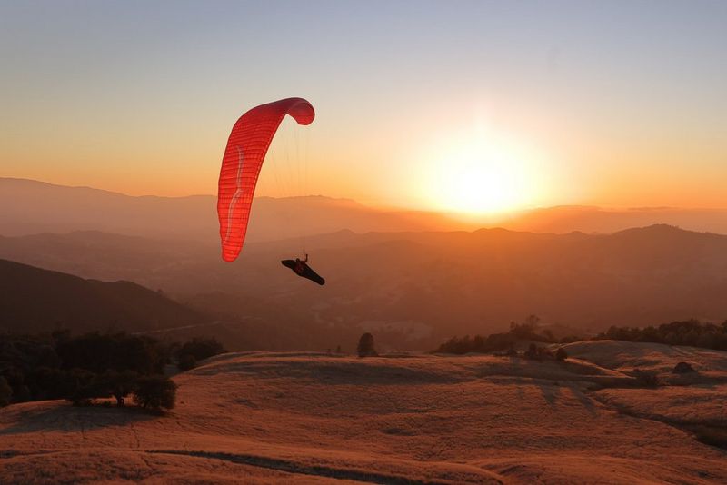 A paraglider with red sail is midair above an arid landscape with a bright sun just above the horizon in the background.