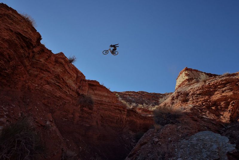 A mountain bike and rider are high in the air jumping between rocks against a rugged landscape and bright blue sky.