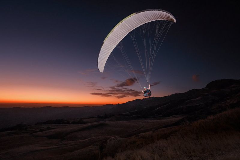 A paraglider with white sail is caught across a twilight sky, the sun just below the horizon. 