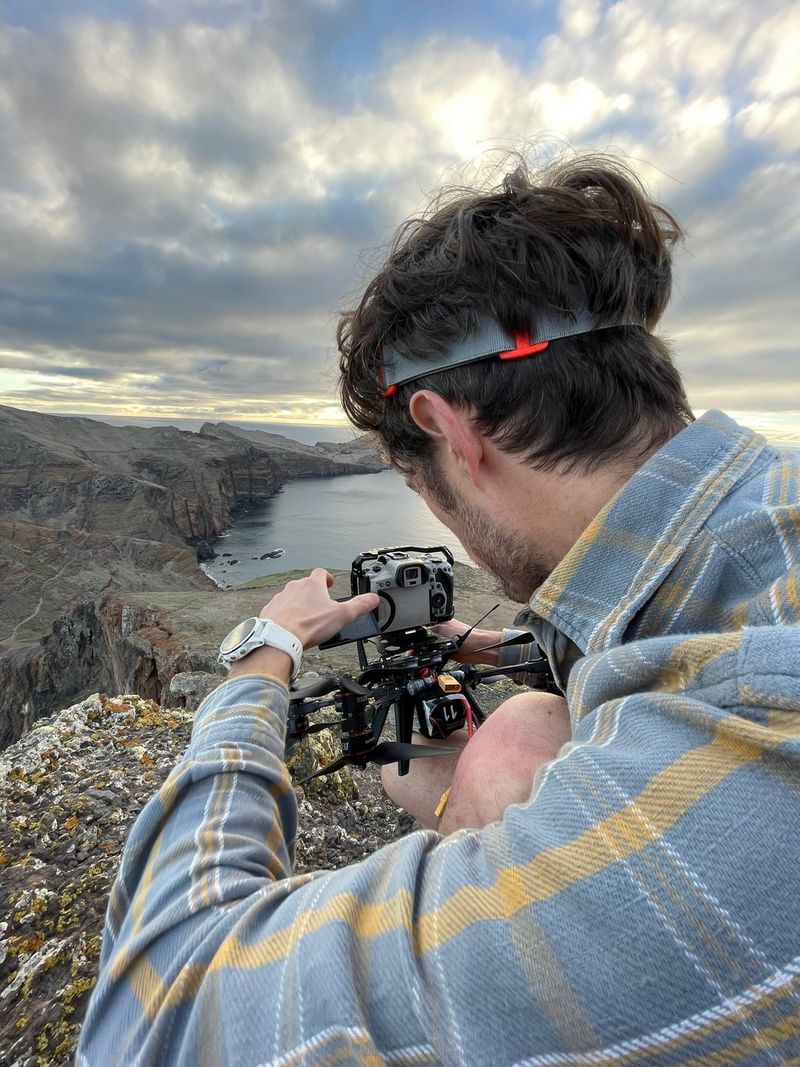 Een opname over de schouder van een man die op de rand van een klif zit en een Canon-camera afstelt die boven op een FPV-drone is bevestigd.