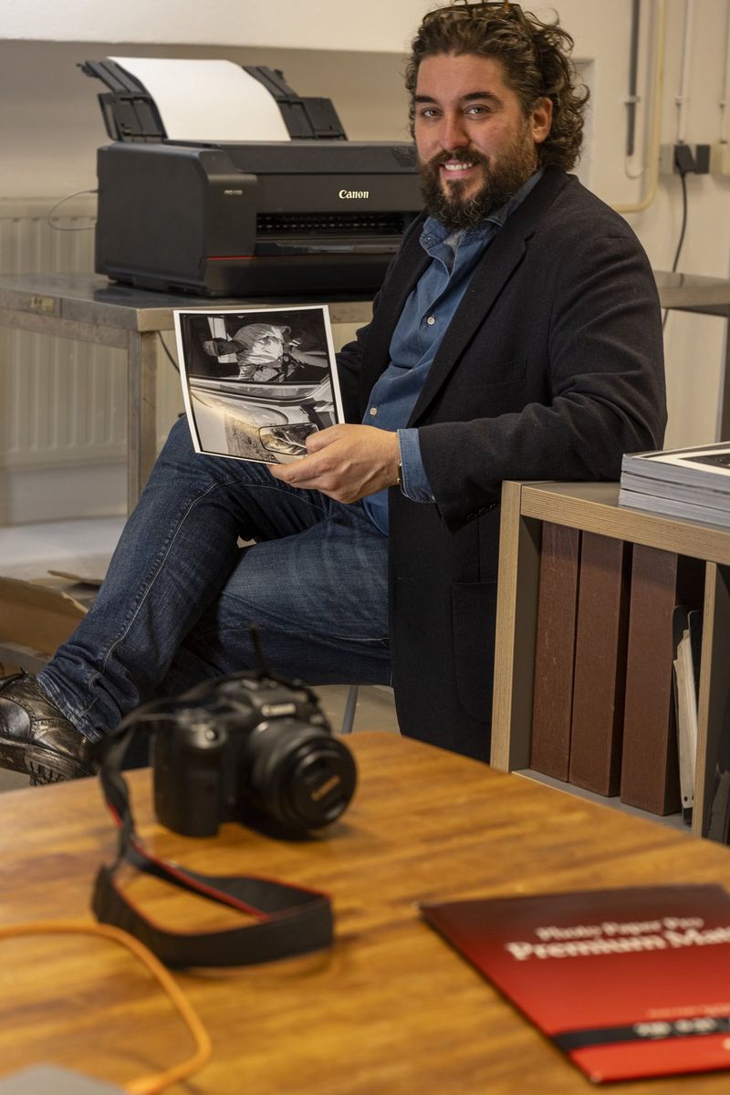 Ahmet Polat sits holding a photo print in front of a Canon imagePROGRAF PRO-1100 printer. On a table in the foreground are a Canon camera and a packet of Canon paper.