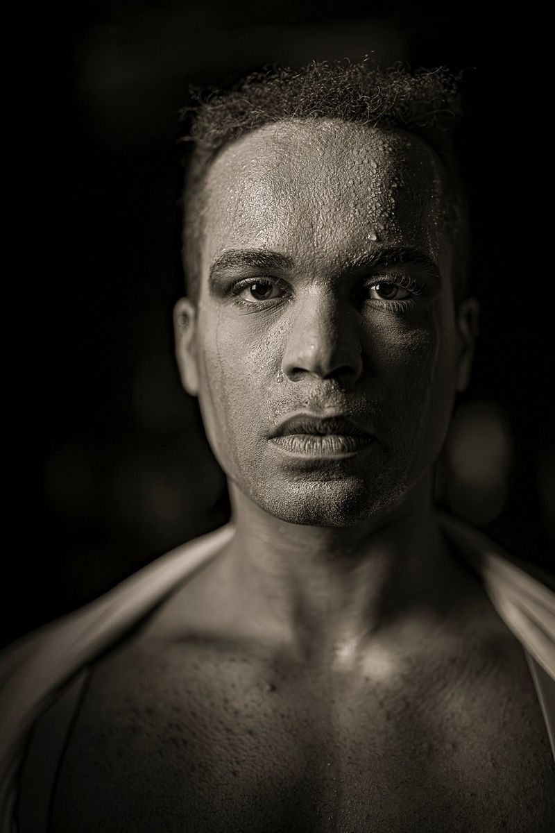 A low-light black and white portrait of a dancer looking straight at the camera, beads of perspiration on his forehead. Taken by Clive Booth on a Canon EOS R5 Mark II with a Canon RF 50mm F1.2L USM lens.   