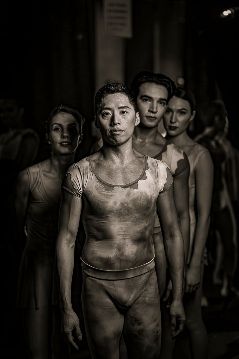 A low-light black and white group shot of dancers standing in the wings with other people looking almost painted in the background. Taken by Clive Booth on a Canon EOS R5 Mark II with a Canon RF 50mm F1.2L USM lens.