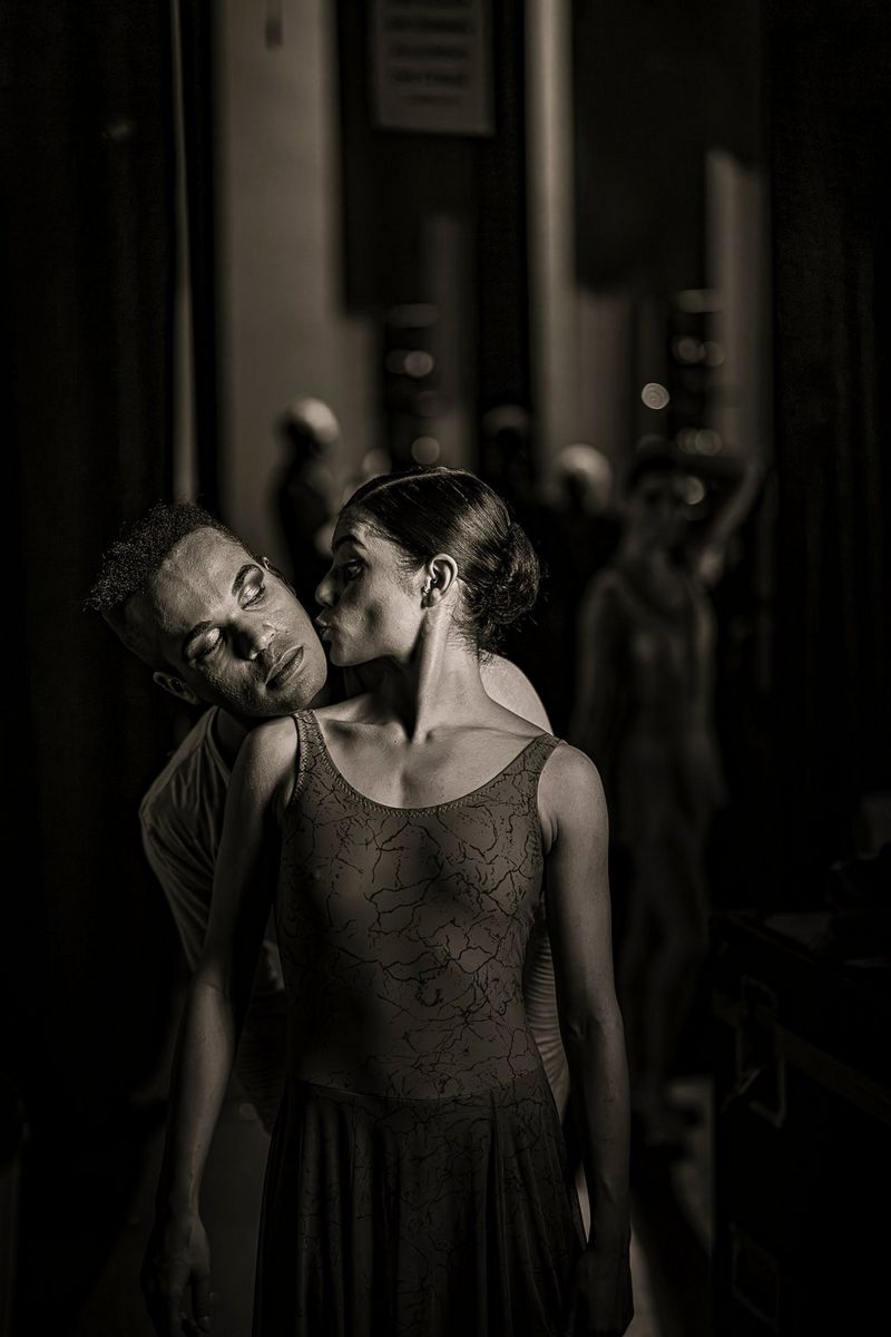A low-light black and white portrait photo of two dancers, one kissing the cheek of the other as he rests his chin on her shoulder from behind. Taken by Clive Booth on a Canon EOS R5 Mark II with a Canon RF 50mm F1.2L USM lens.