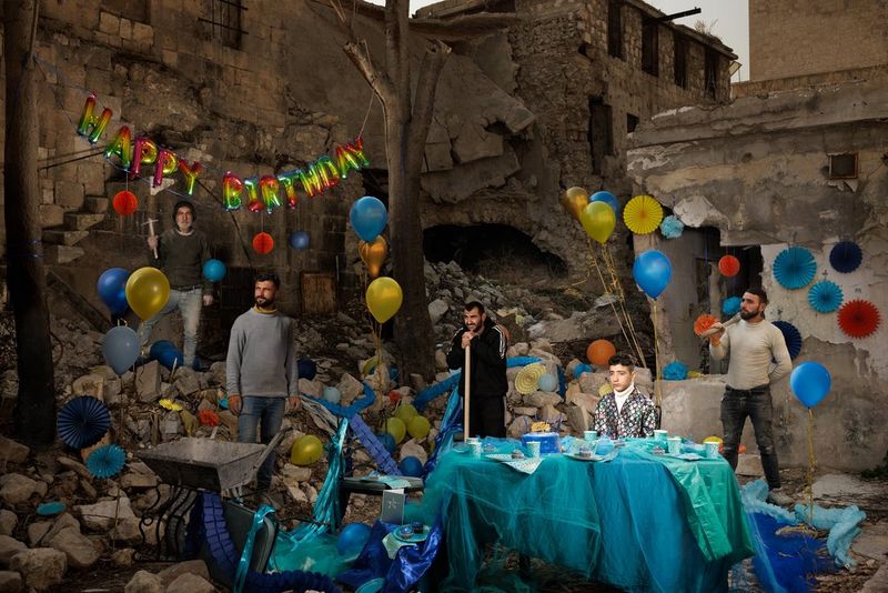A young boy sits at a party table draped in blue, with coloured balloons and a banner spelling out the words “happy birthday” in the background. All around is rubble and ruined buildings, and other people in the picture stand with a wheelbarrow, picks and other tools. Fantasy portrait taken by Elisa Iannacone on a Canon EOS R5 C with a Canon RF 24-70mm F2.8L IS USM lens.