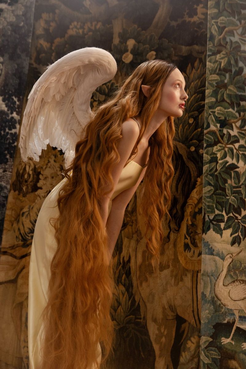 Against a backdrop like a pre-Raphaelite painting, a model made up like a fairy, with pointed ears and fluffy white wings, leans forward, her long auburn hair cascading almost to her knees. Fantasy portrait taken by Wanda Martin.