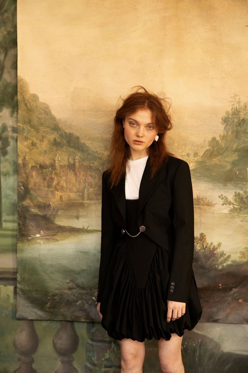 A model in a black jacket and pleated skirt stands in front of a painted landscape backdrop. Fashion photo taken by Wanda Martin.