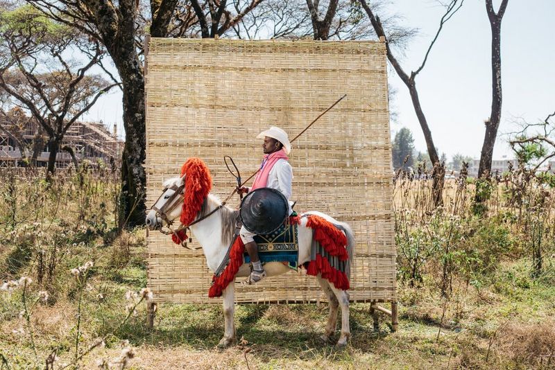 Una persona seduta su uno sgabello davanti a un separé di paglia, in una fotografia scattata con Canon EOS 5D Mark IV da Mekbib Tadesse.
